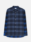Flat lay of a Universal Works navy and blue brushed flannel check shirt on a light grey studio backdrop. The body features a navy and slate check while the sleeves and hem panel use a contrasting bright blue, black and navy tartan. Point collar with button-down tabs, white buttons down the placket, a chest patch pocket, and a curved shirttail hem.