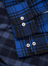 Close-up detail of a navy and blue brushed flannel check shirt. The cuff features two white buttons and a rounded placket edge, with visible topstitching. A chest patch pocket with a curved flap and single white button sits above. The tartan-style check pattern shows contrasting tones across panels. No backdrop visible.