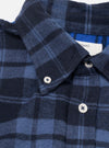 Close-up of a navy and blue brushed flannel check shirt on a white studio backdrop. The shot focuses on the collar and button placket, showing a button-down collar fastened with a white button. Two white buttons are visible on the front placket. A woven label is partially visible at the collar. No model or mannequin present.