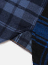 Close-up detail of a navy and blue brushed flannel check shirt, showing the hem and side seam where two contrasting check panels meet. The left panel features a tonal navy check and the right a bold blue and black check. Orange topstitching runs along the curved hem. White studio backdrop.