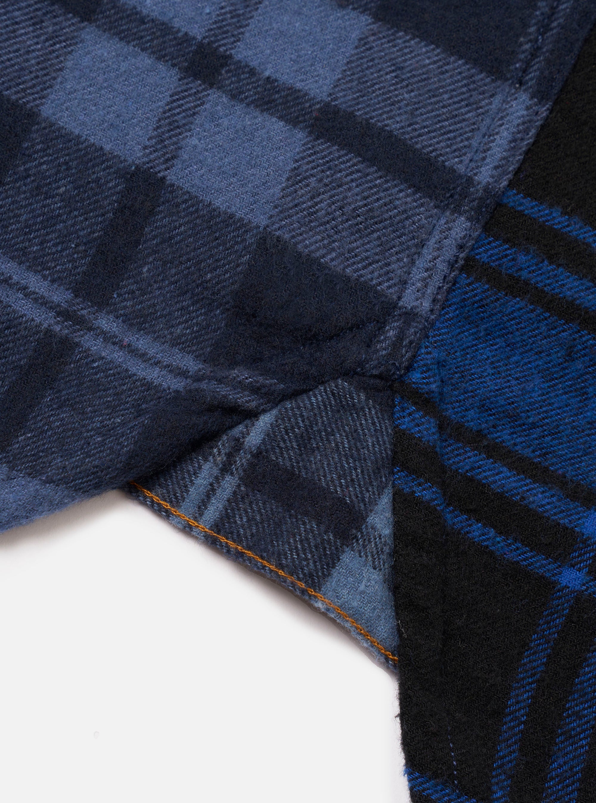 Close-up detail of a navy and blue brushed flannel check shirt, showing the hem and side seam where two contrasting check panels meet. The left panel features a tonal navy check and the right a bold blue and black check. Orange topstitching runs along the curved hem. White studio backdrop.