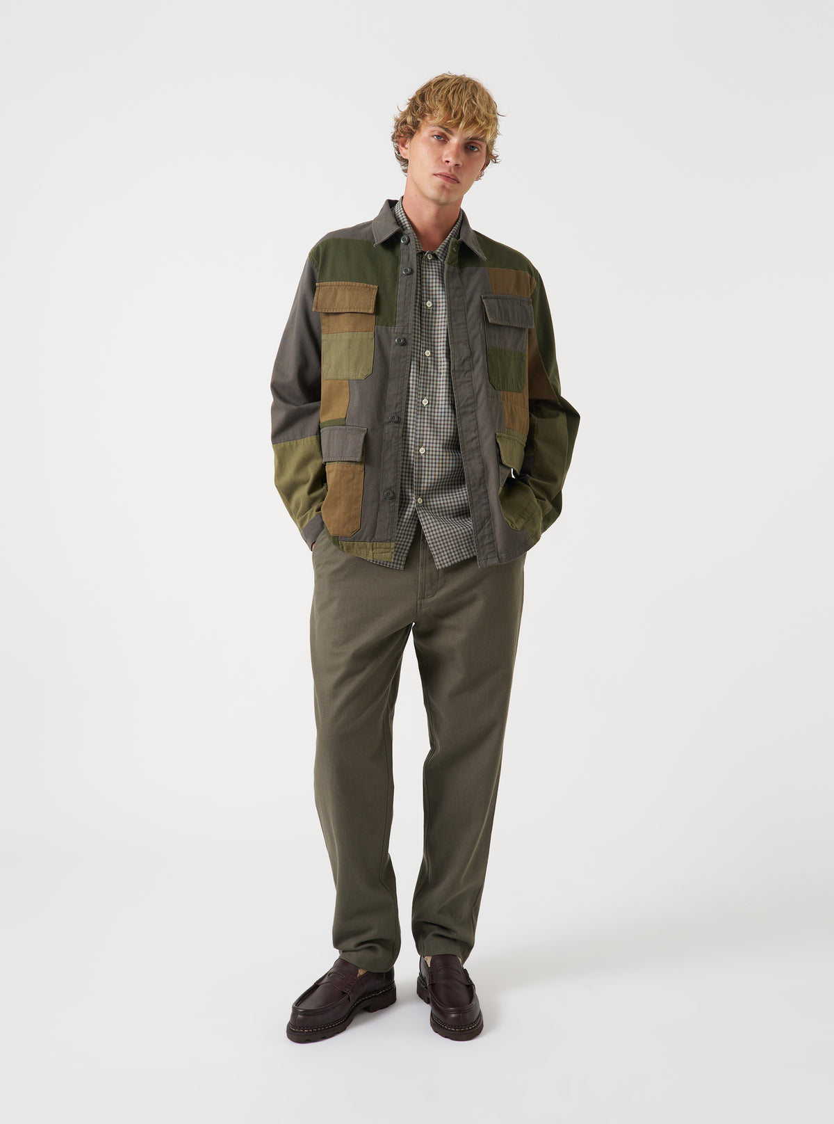 A full-length studio shot of a model wearing the Olive Patchwork Twill MW Fatigue Jacket against a neutral background. The photo showcases the military jacket styling, front button closure, flap pockets, and patchwork twill fabric.