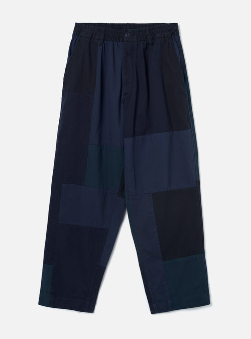 Navy patchwork twill Oxford trousers laid flat on a plain light grey backdrop. Wide-leg cut with an elasticated waistband, single centre button, and slant side pockets. The body features large irregular patchwork panels in varying shades of navy, teal and black woven twill. Full-length leg with an unfinished hem.