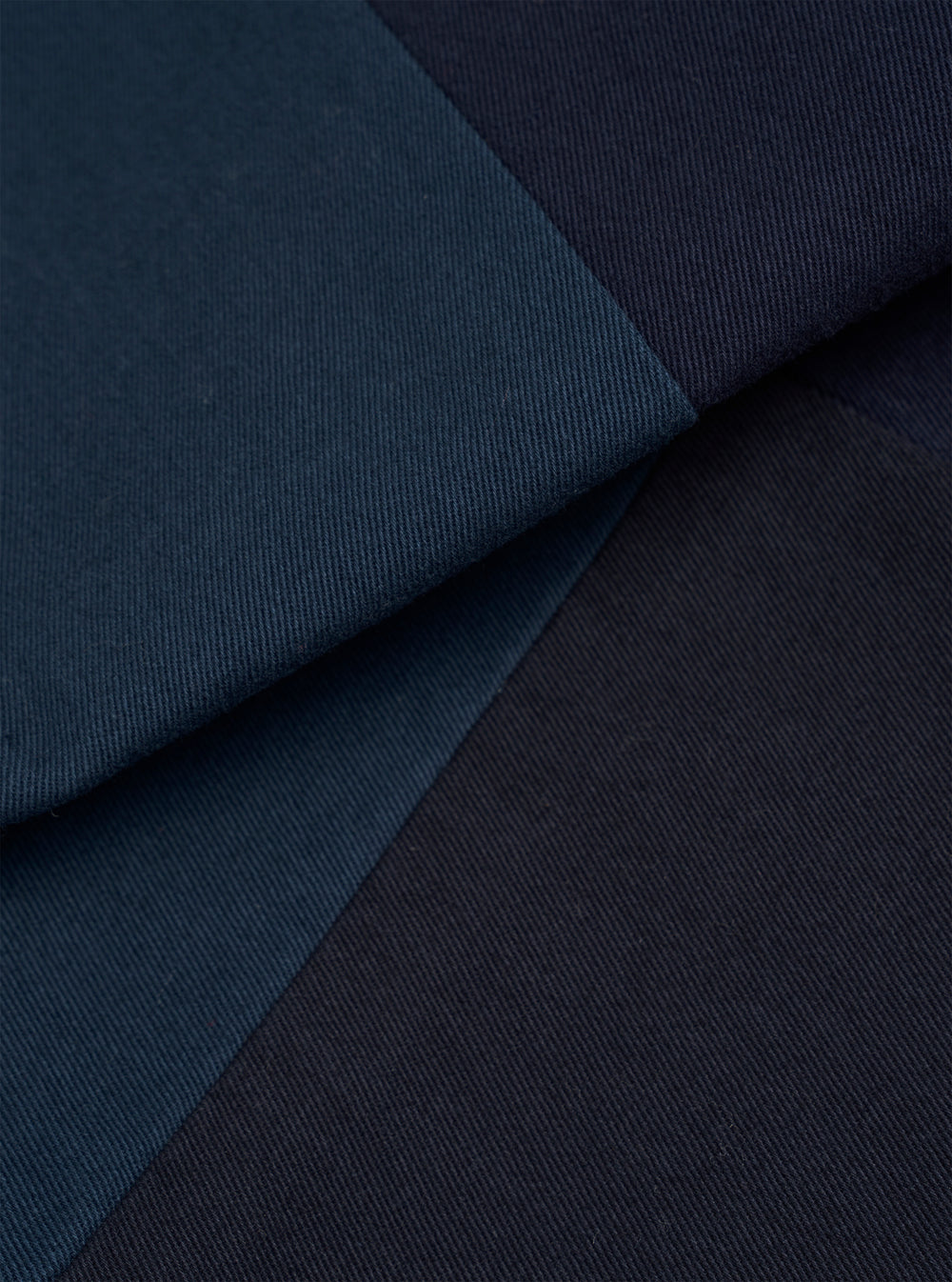 Close-up detail of navy twill fabric showing multiple folded layers in varying shades of navy, from mid-blue to deep navy. The fine diagonal twill weave is clearly visible across all panels. Shot against a plain dark background with no model or mannequin present.