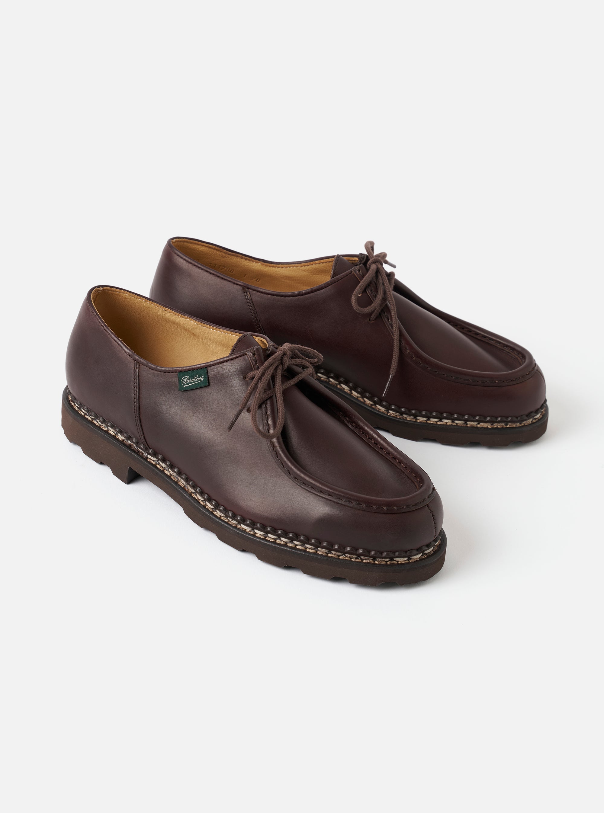 Pair of dark brown leather Paraboot Michael Marche II shoes shot on a light grey studio backdrop. Both shoes display a moc-toe apron seam, visible welt stitching in contrasting cream thread, a chunky dark rubber lug sole, and brown waxed laces. A small green Paraboot branded label is visible on the left shoe. Tan leather lining is visible at the collar.