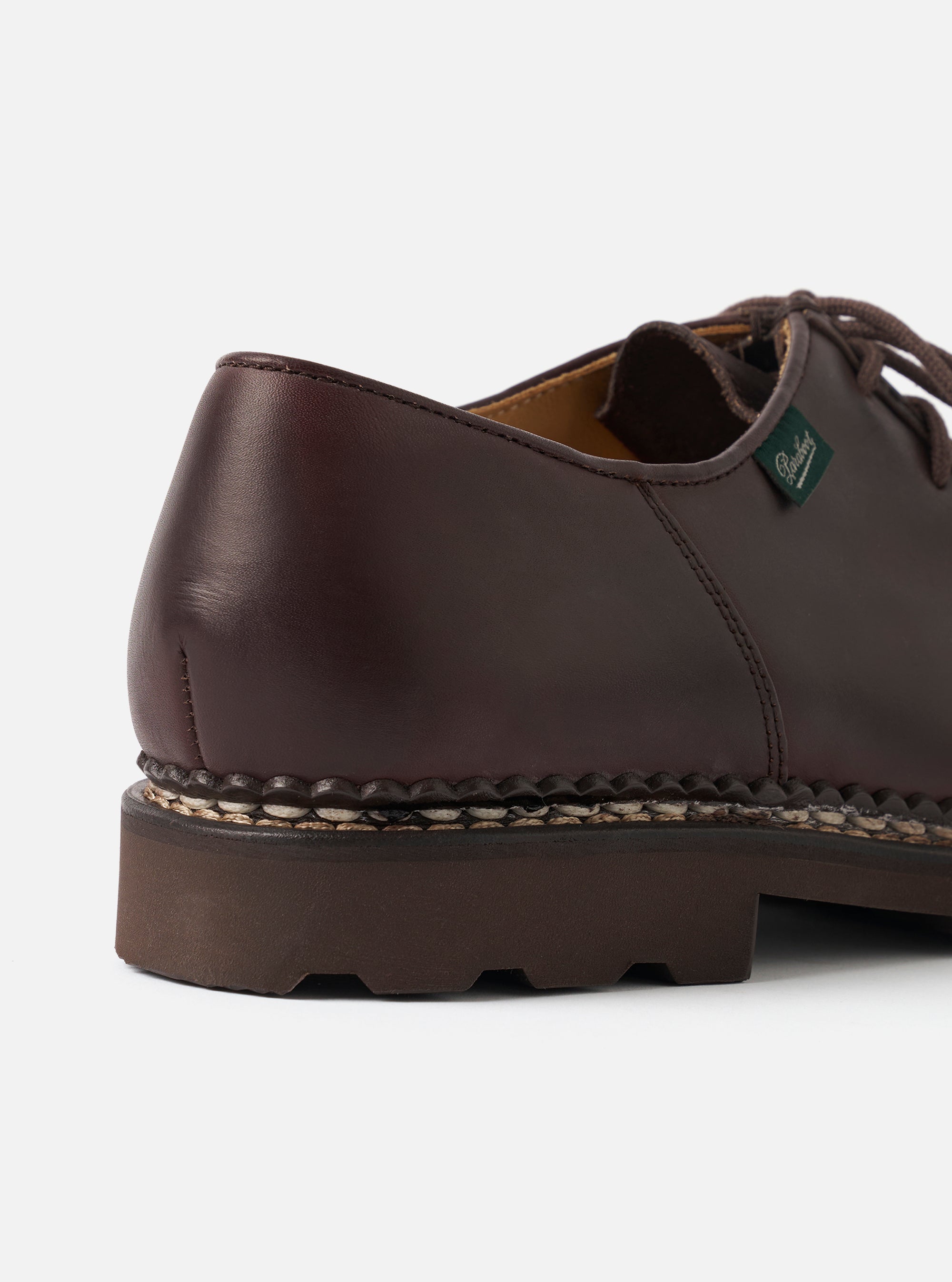 Close-up of the heel and quarter of a Paraboot Michael Marche II in cafe brown leather. Smooth, polished leather upper with a green Paraboot branded tongue tag visible. Distinctive Norwegian welt stitching runs along the midsole join. Chunky dark brown rubber sole with a lugged profile. Plain light grey studio backdrop.