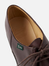 Close-up of a Paraboot cafe brown leather derby shoe on a light grey studio backdrop. The tongue is folded back to reveal the tan leather insole, embossed with the Paraboot logo and "Cousu Norvégien, Made in France." A green Paraboot branded label is visible on the tongue. Round brown laces are partially visible.