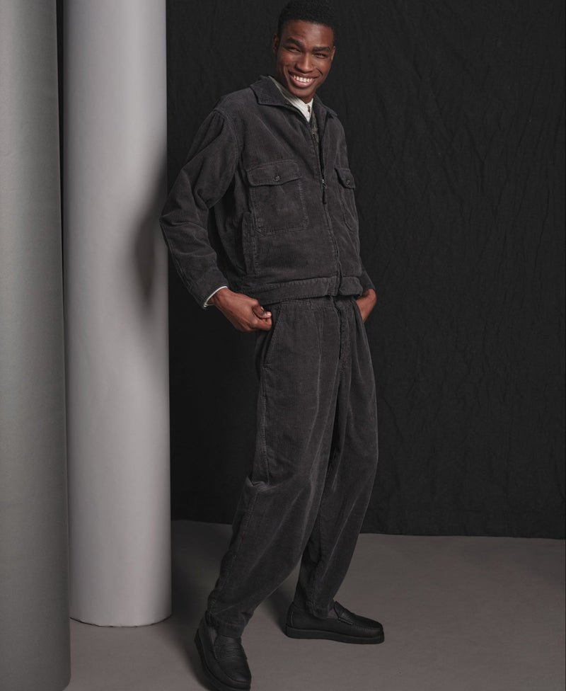 Man wearing a dark gray corduroy matching jacket and trousers standing against a black background