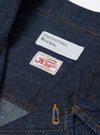 The Indigo Kaihara LT WT Denim Jungle Jacket is shown in a close-up detail shot focusing on the interior neck area against a white background. The image prominently features the Universal Works brand label sewn onto the indigo denim fabric, alongside a smaller white size tag. The silver metal hardware of a top button is visible, and the collar's construction and stitching are clearly shown.