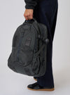 A person holds the F/CE.® Grey Recycled Nylon 420 re/cor Travel Backpack. The grey nylon backpack is held against a white backdrop, with a person wearing dark blue pants and black sneakers.