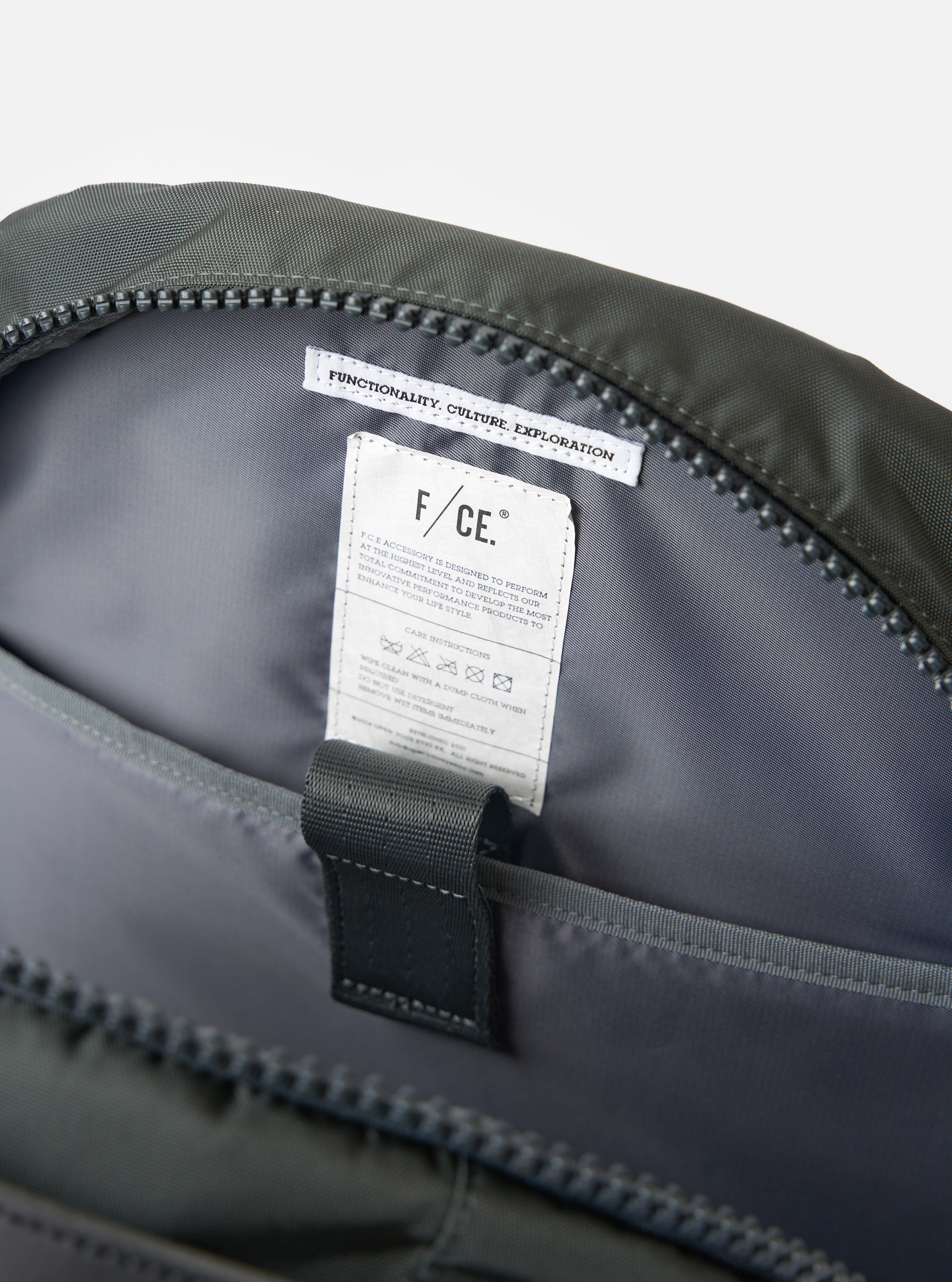 Close-up of the inside of the F/CE.® Grey Recycled Nylon 420 re/cor Travel Backpack. Shows the grey nylon lining, zipper, and care instructions tag. Shot on a white background.