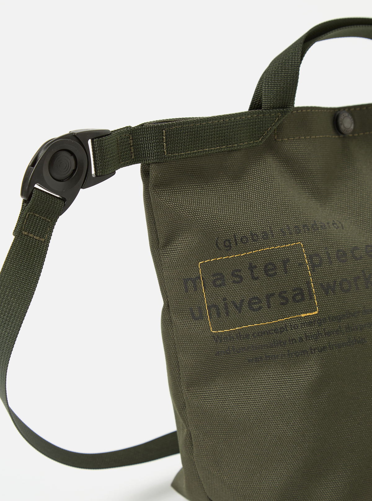 Close-up of the master-piece x Universal Works Olive Recycled Tech Canvas Small Tote Bag. The olive green bag features black lettering and straps, set against a clean white background.