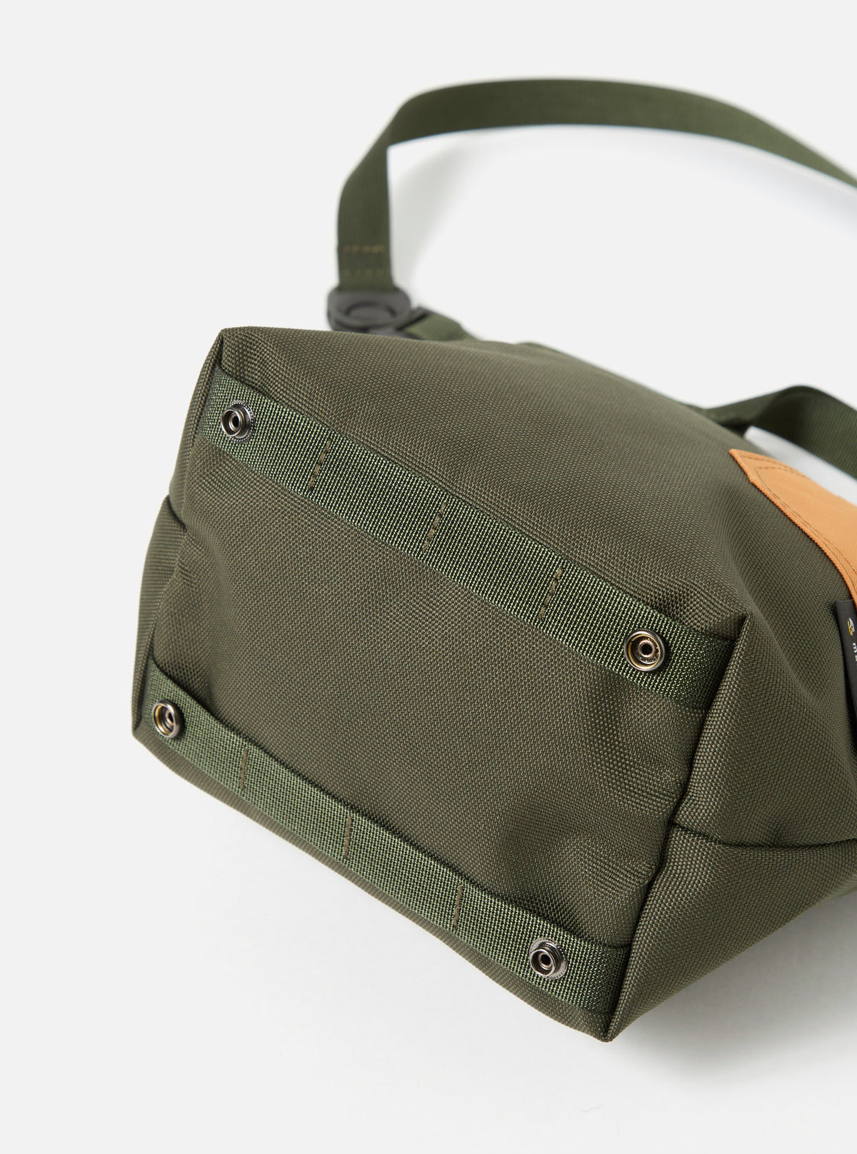 Close-up of the base of a master-piece x Universal Works Olive Recycled Tech Canvas Small Tote Bag on a white surface. The olive green canvas is detailed with webbing and metal rivets.
