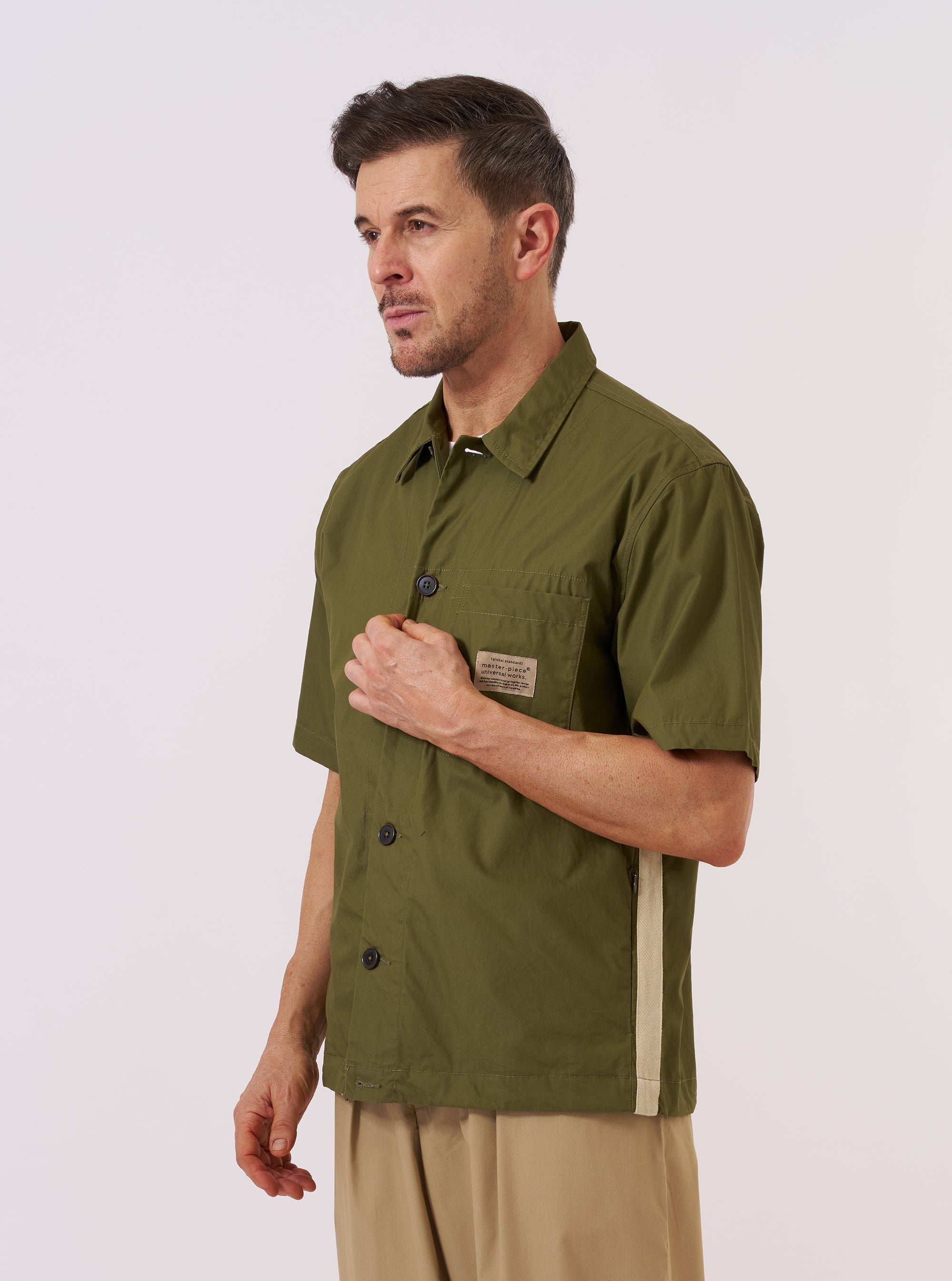 A model wears the master-piece x Universal Works Olive Recycled Poly Tech Tech Overshirt against a plain white background. The olive green overshirt features short sleeves, a one-piece collar, a central button-down closure, and a single patch pocket on the left chest. The model is shown from the front, with the garment displaying a clean, technical finish and a straight hem.