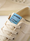 The Novesta for Universal Works Ecru/Pale Blue Canvas Star Master is shown from a side profile view against a white background. The sneaker features an ecru cotton canvas upper with matching ecru laces and metallic eyelets. A prominent feature is the thick, vulcanized natural rubber sole in ecru with a textured toe cap. A signature pale blue fabric heel pull tab is visible at the back, and a dual-branded woven logo tag is attached to the tongue.