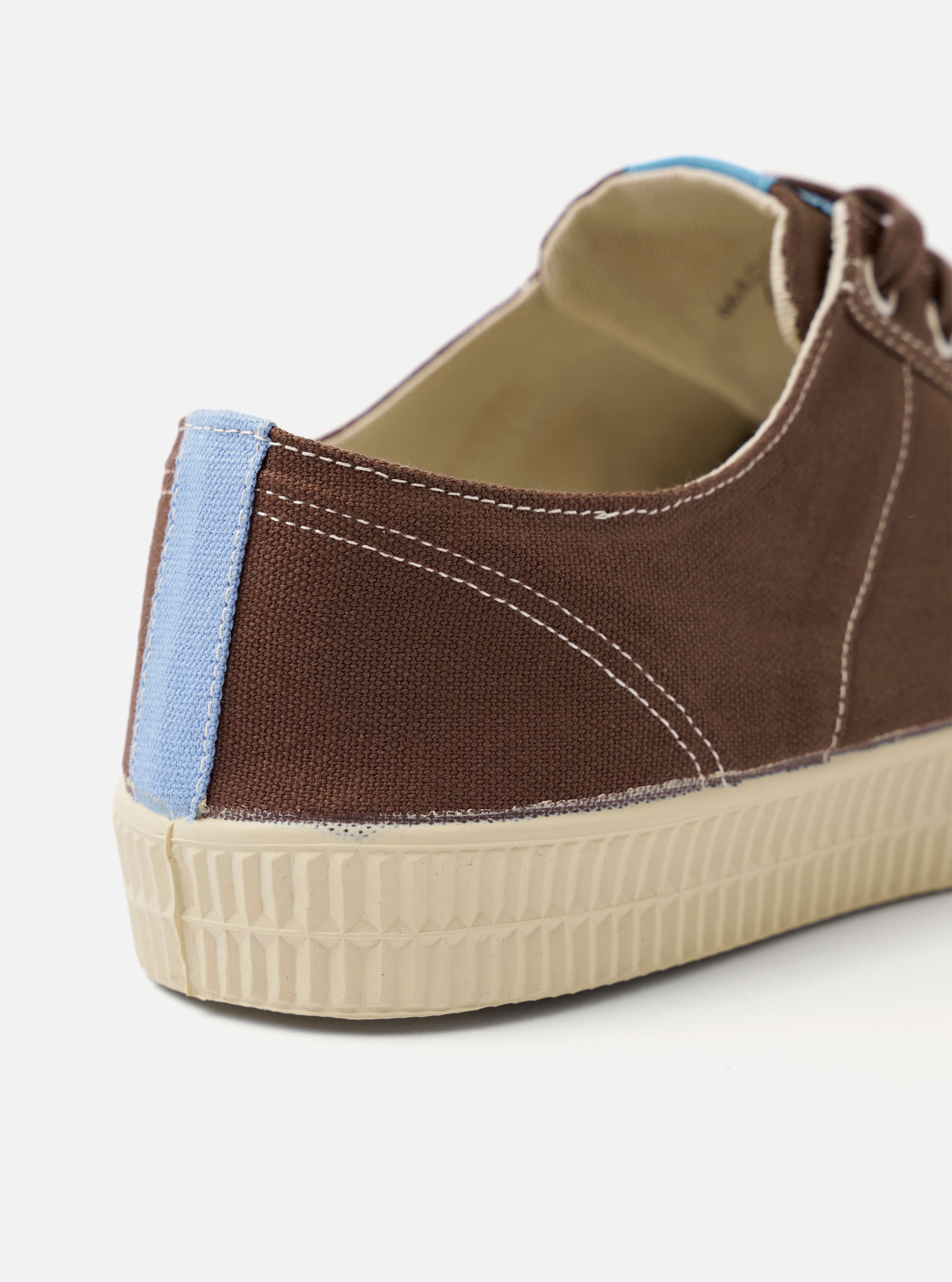 The Novesta for Universal Works Brown/Pale Blue Canvas Star Master shoe is shown from a side profile view against a plain white background. The sneaker features a brown cotton canvas upper with contrast stitching, metallic eyelets, and a chunky, textured ecru vulcanized rubber sole. A distinctive pale blue pull tab is located on the heel, and a dual-branded woven tag is visible on the tongue.