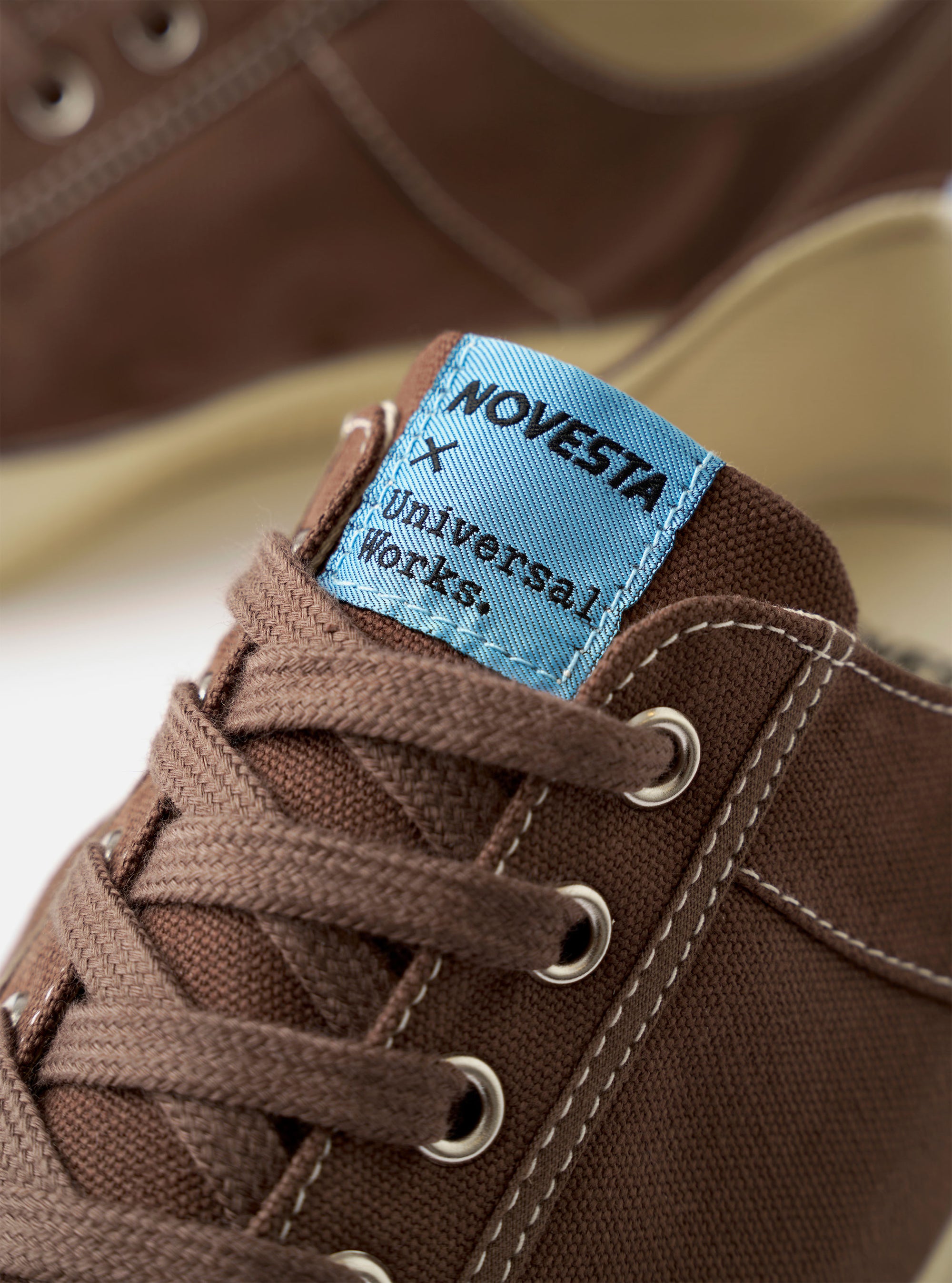 The Novesta for Universal Works Brown/Pale Blue Canvas Star Master sneaker is shown from a side profile against a white background. The shoe features a dark brown cotton canvas upper with contrast stitching, silver metallic eyelets, and a chunky ecru-colored vulcanized rubber sole with a textured toe cap. A distinctive pale blue pull tab is visible on the heel, and a dual-branded woven tag is stitched onto the tongue.