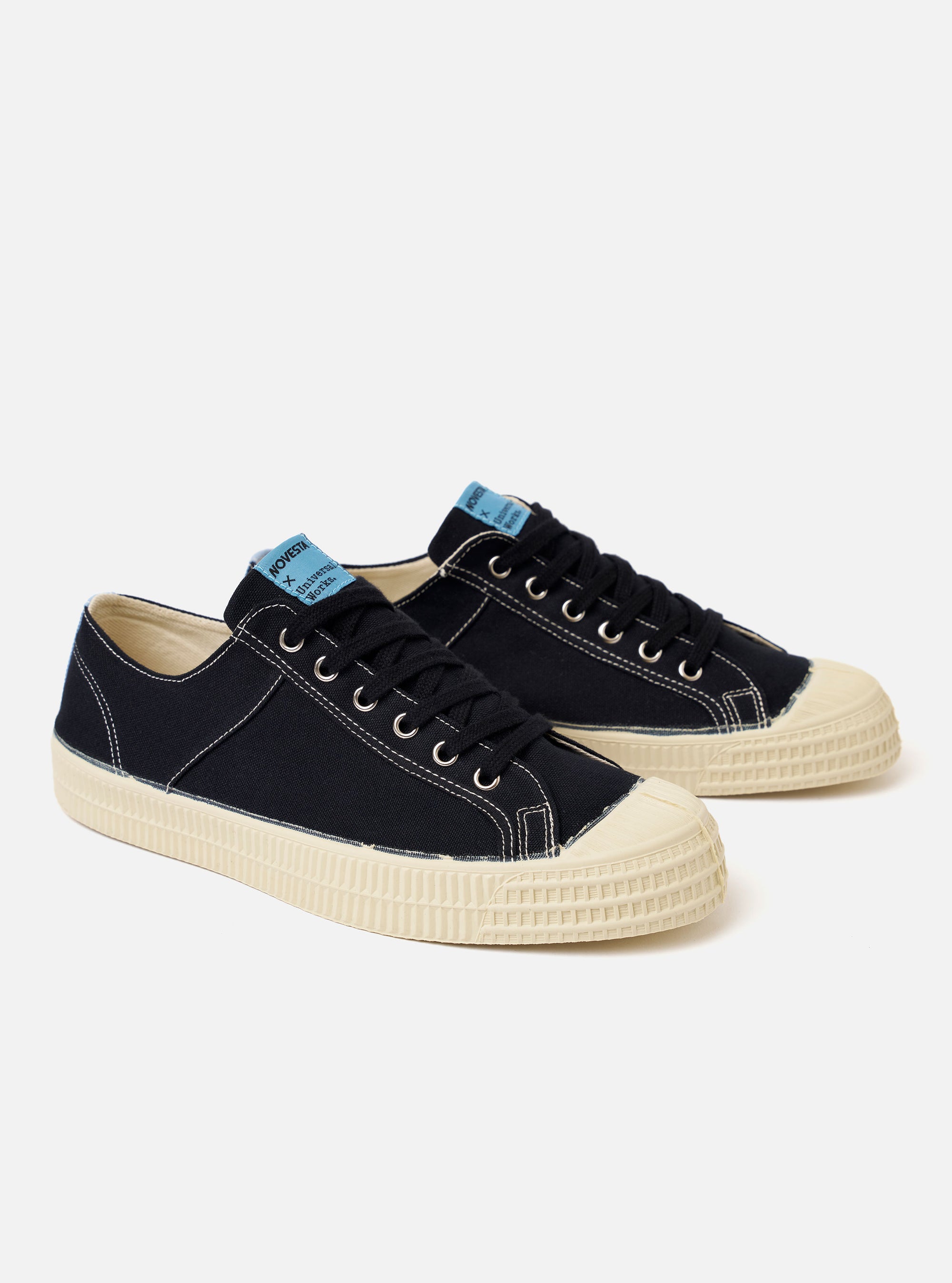 The Novesta for Universal Works Black/Pale Blue Canvas Star Master is shown from a high-angle, side profile view against a white background. The sneaker features a black cotton canvas upper with contrast stitching, metallic eyelets, and a chunky ecru vulcanized rubber sole with a textured toe cap. A pale blue pull tab is visible on the heel, and a dual-branded woven tag is on the tongue.