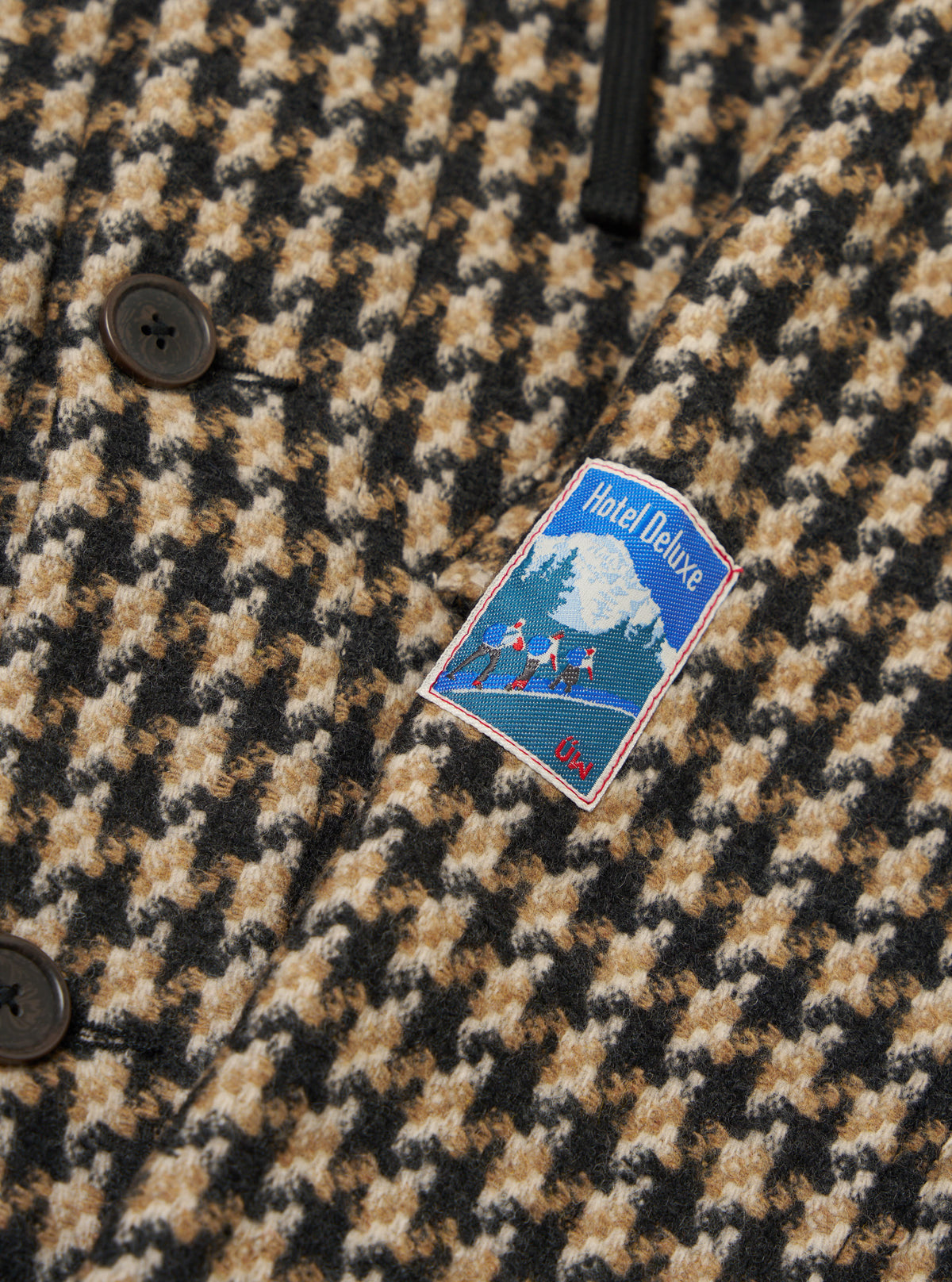 Close-up of the Sand Cortina Tweed Boston Parka showing the tweed fabric, buttons, and Hotel Deluxe patch. The background is a plain surface.