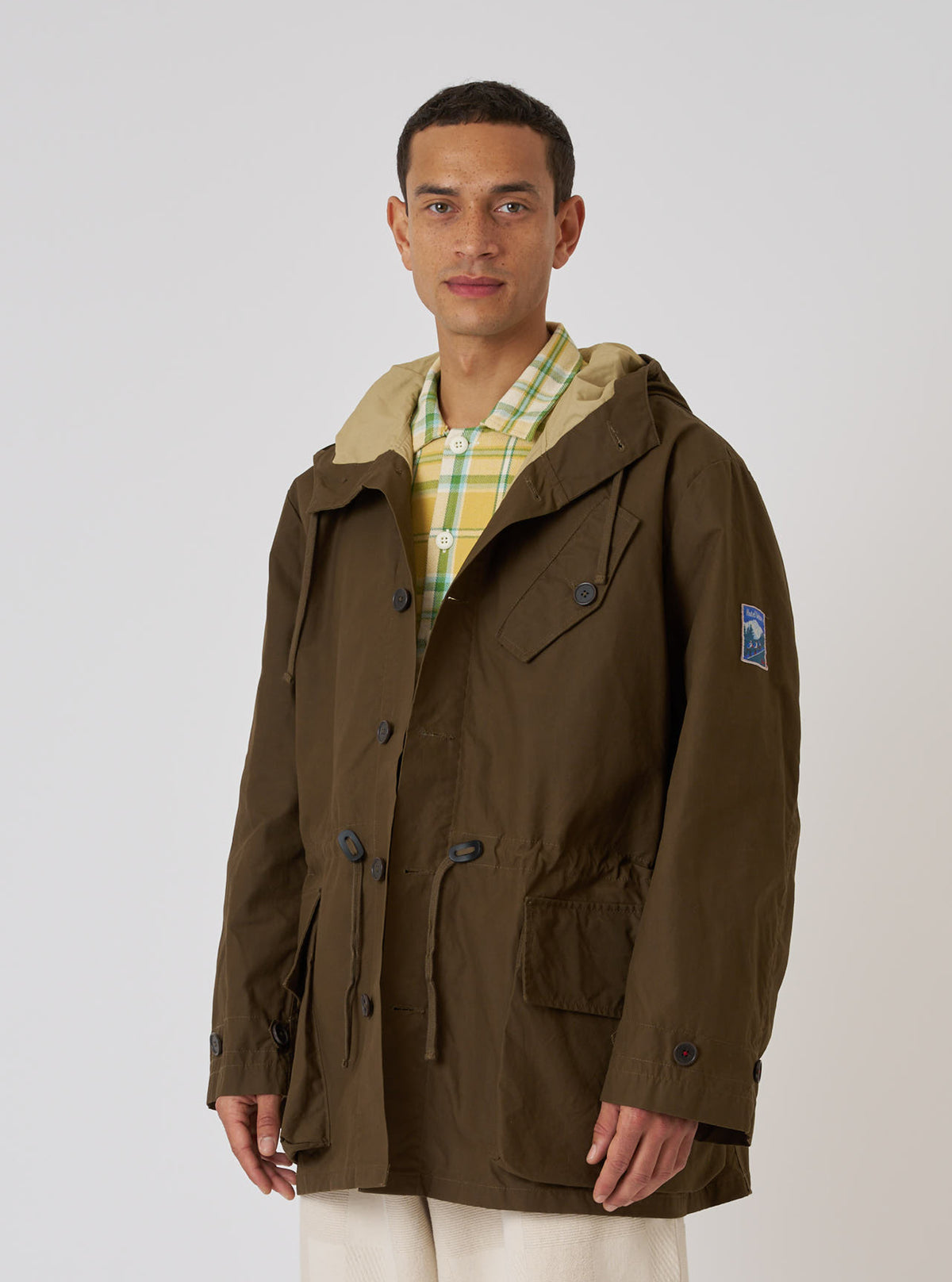A model wears a Brown British Wax Cotton Boston Parka in front of a white background. The parka is brown and has a hood and pockets.