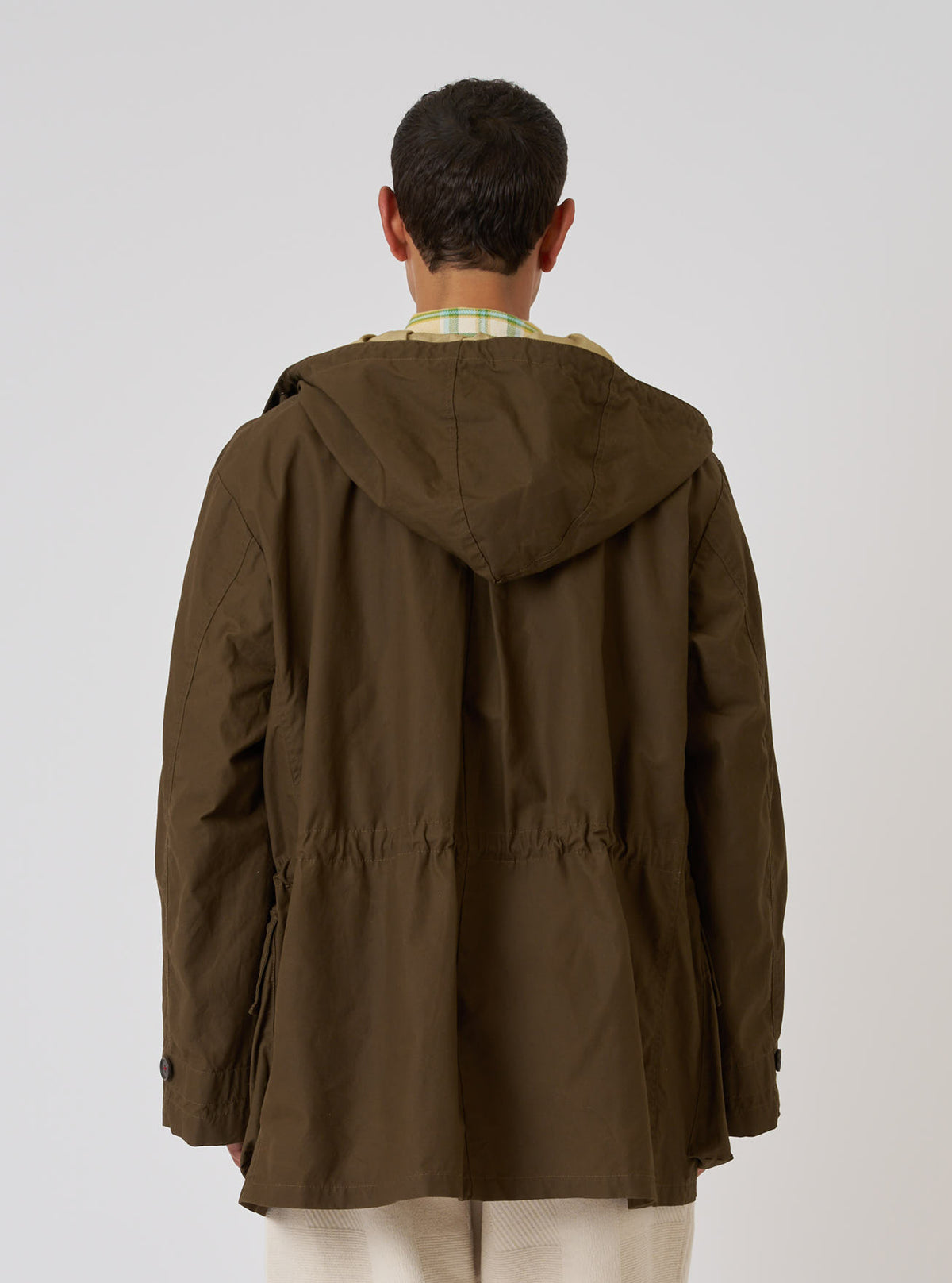 Back view of a model wearing a Brown British Wax Cotton Boston Parka. The parka is brown and made of wax cotton. The background is a plain white studio backdrop.