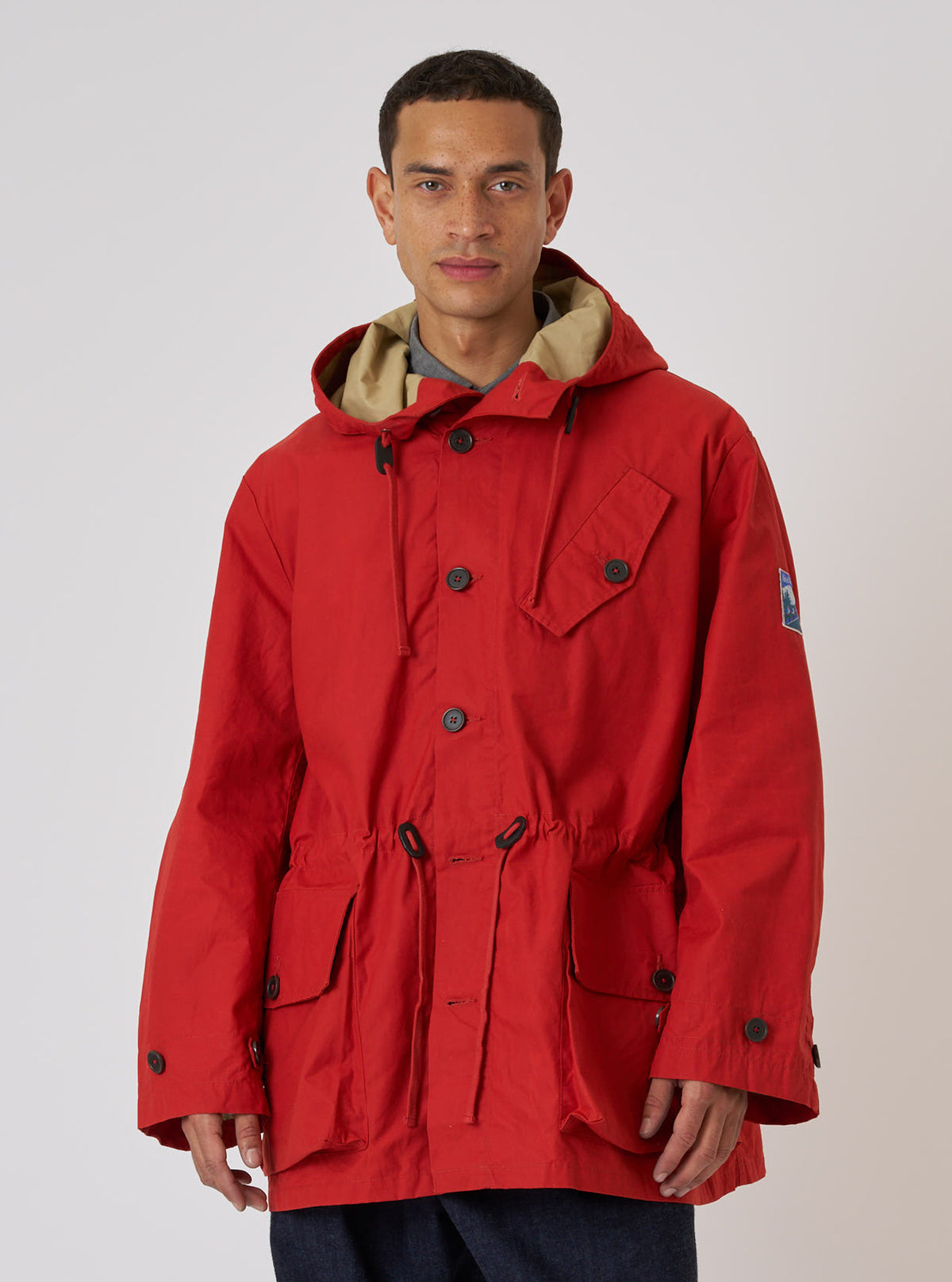 A model poses in an Orange British Wax Cotton Boston Parka against a white backdrop. The parka features a tan hood lining, button closure, and drawstring waist.