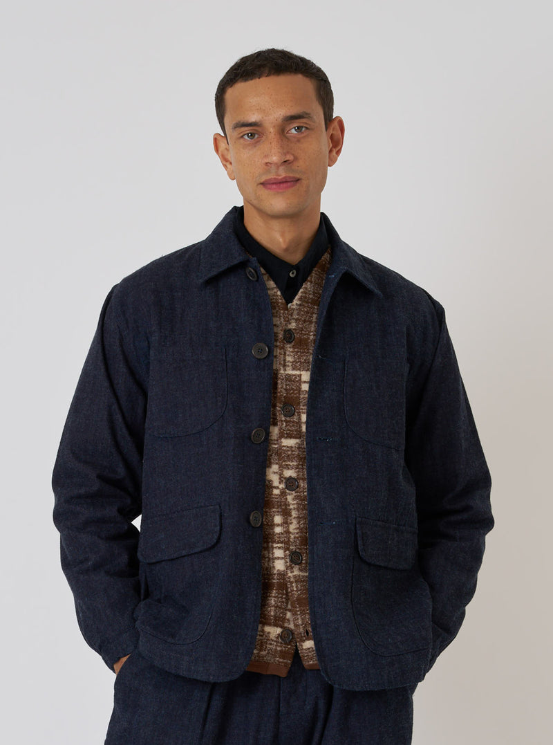 A model poses in an Indigo Brushed Denim Labour Jacket against a white backdrop. The jacket is paired with a patterned vest and dark shirt, showcasing its versatile fit.