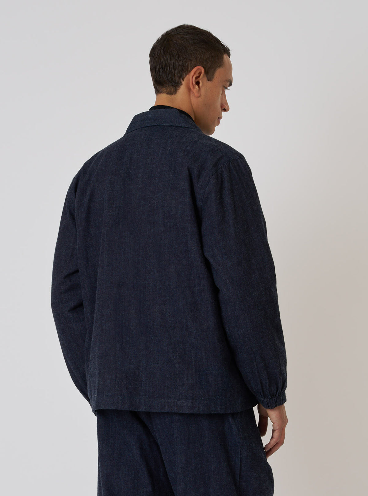 A man poses in the Indigo Brushed Denim Labour Jacket against a plain background. The jacket is a dark indigo color and appears to be made of denim. The fit is relaxed.