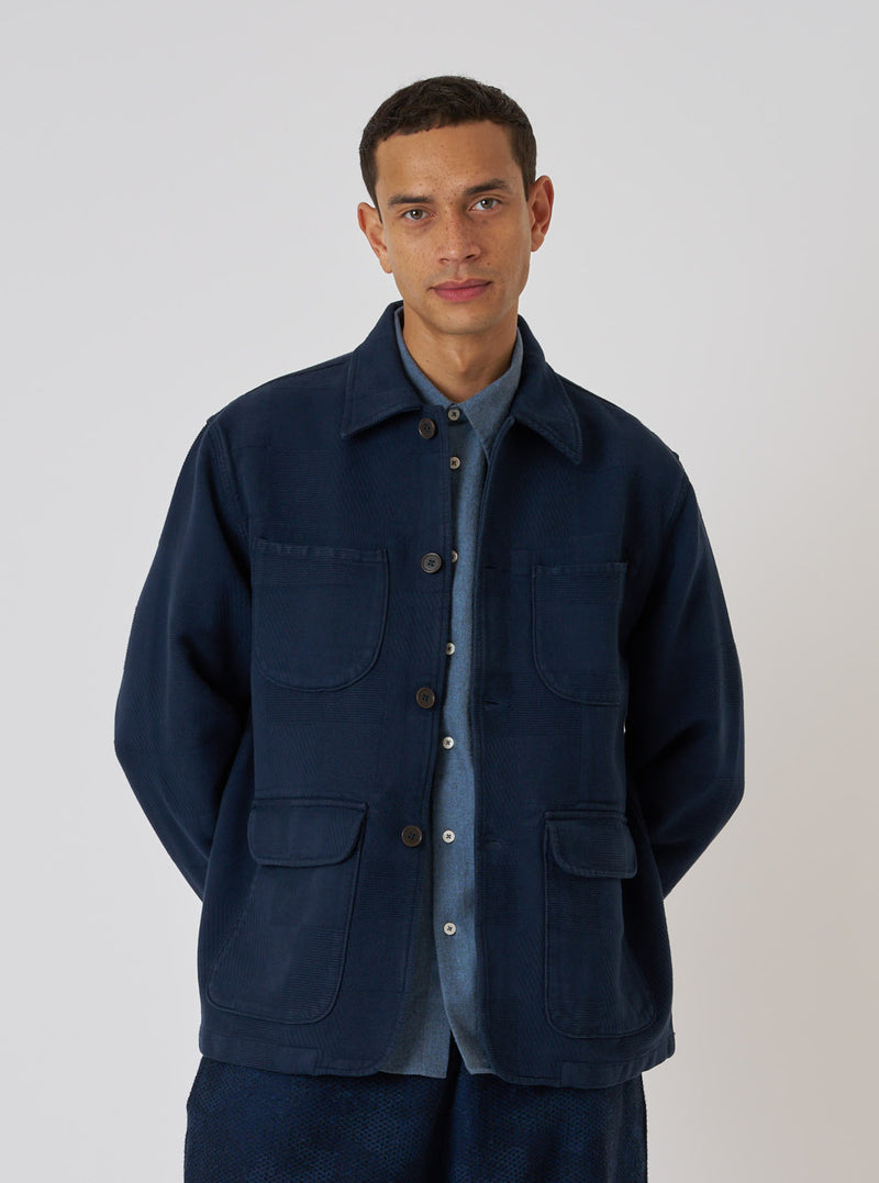 A man poses against a white backdrop wearing a Dark Navy Patchwork Weave Cotton Labour Jacket. The jacket has four pockets and is buttoned over a blue shirt.