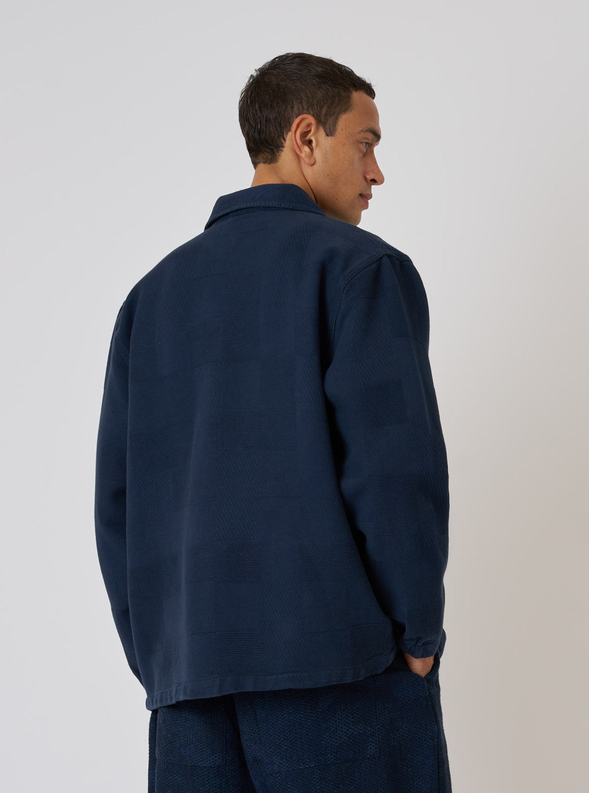 A man models the Dark Navy Patchwork Weave Cotton Labour Jacket against a plain backdrop. The jacket is a dark navy with a subtle patchwork weave.