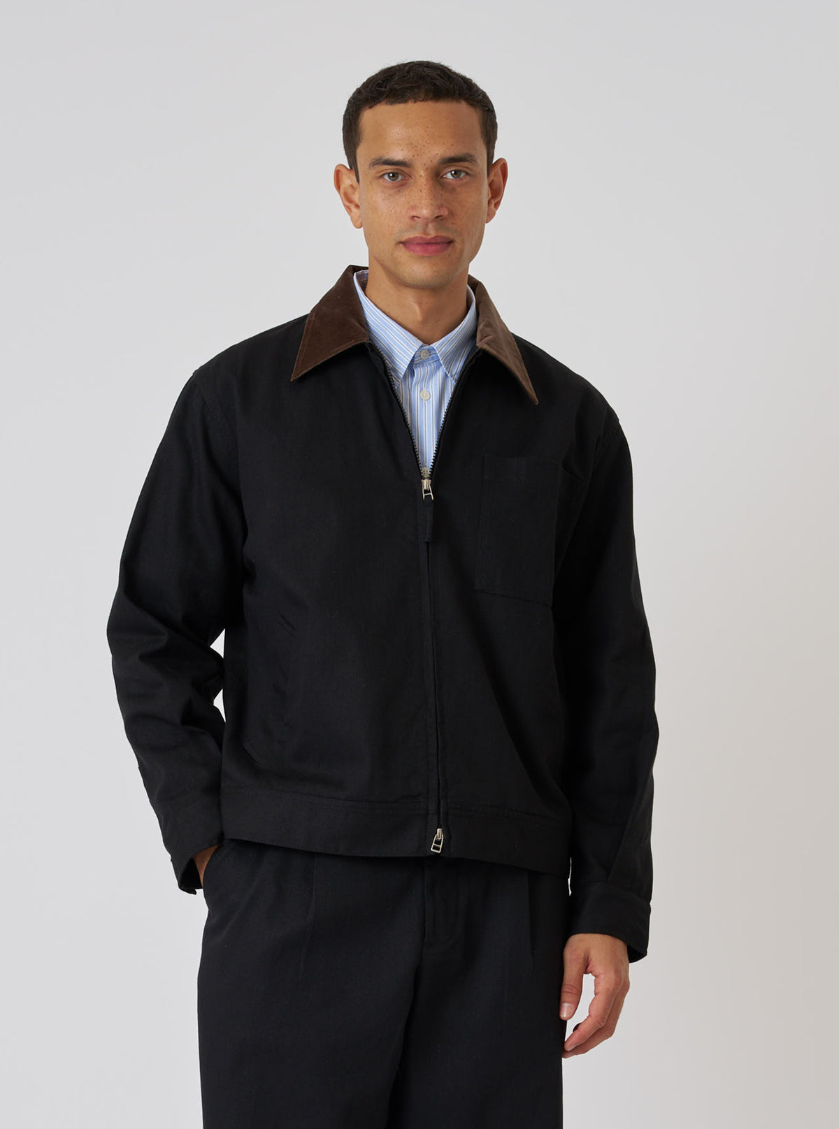 A model poses in a Black Works Twill Rose Bowl Jacket. The jacket is black twill with a brown corduroy collar. The background is a plain white studio backdrop.