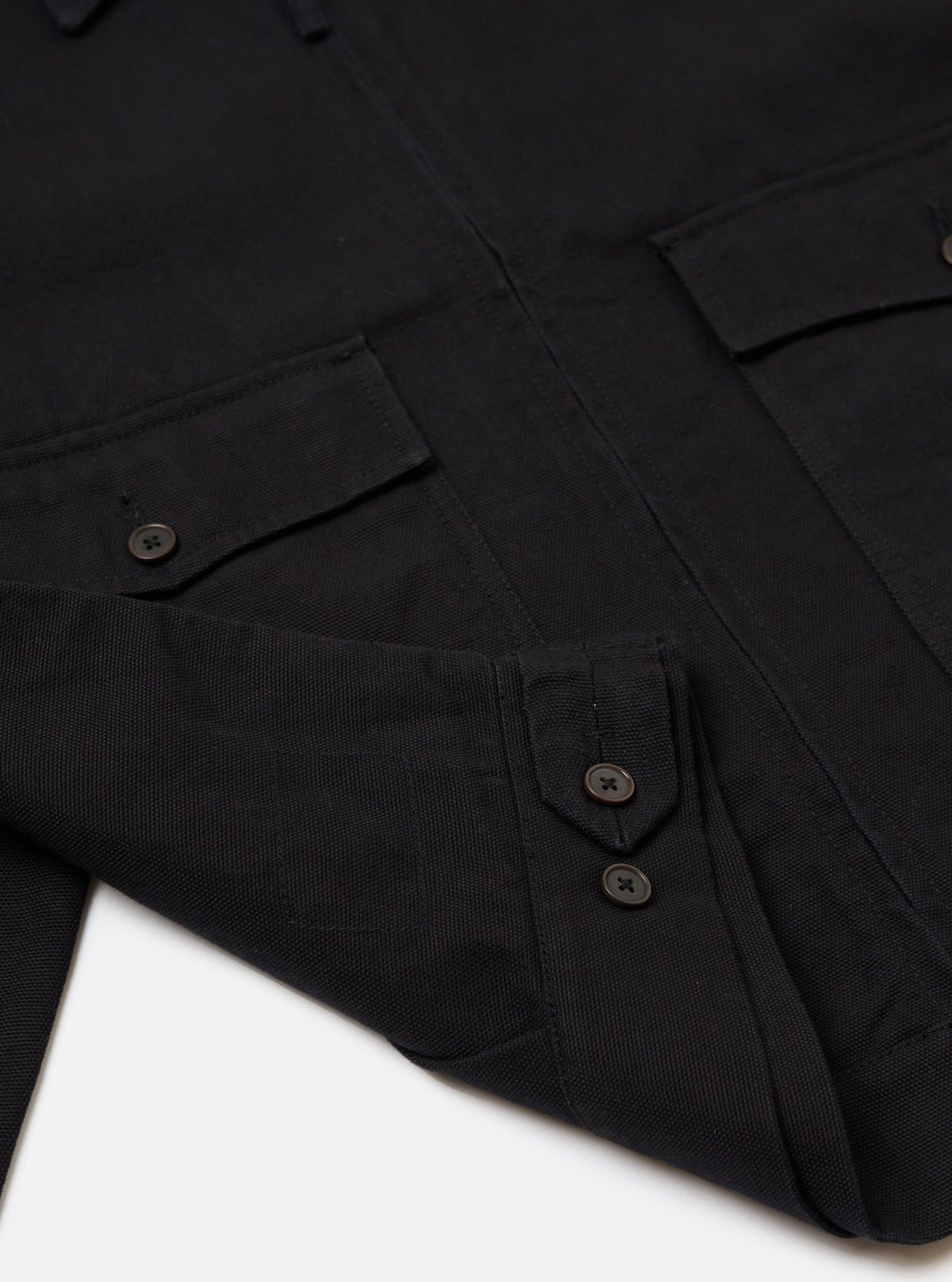 Close-up of the Black Organic Canvas S130 Jacket on a white surface. The jacket features a button-up design and visible pockets, showcasing its organic canvas material.