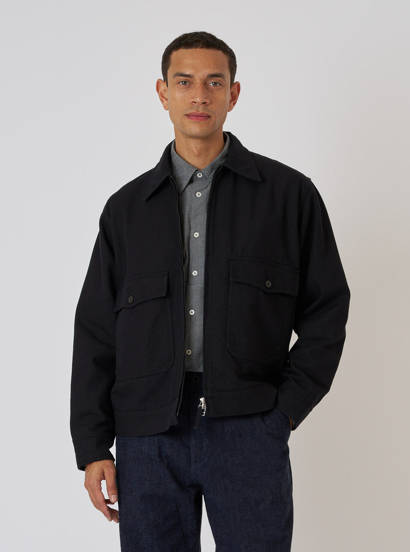 A man poses in front of a white background wearing the Black Organic Canvas S130 Jacket. The jacket is black with a collared neck and two front pockets.