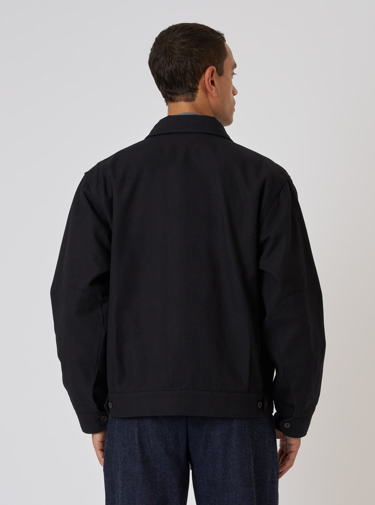 Back view of a man wearing a Black Organic Canvas S130 Jacket. The jacket is black canvas. Shot in a studio setting on a white background.
