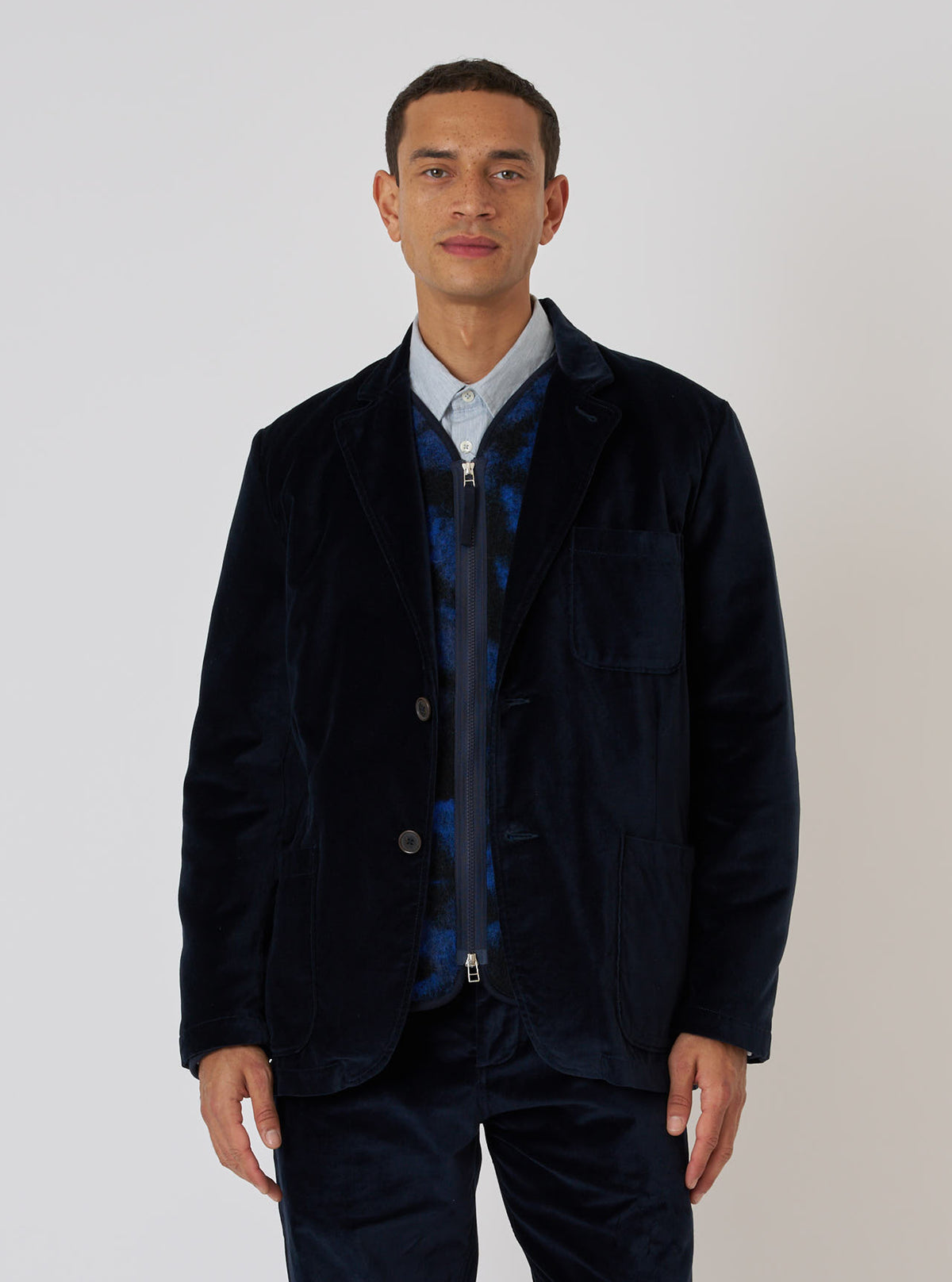 A model poses in front of a white background wearing a Navy Velveteen Two Button Jacket. The jacket is paired with a blue shirt and patterned vest.