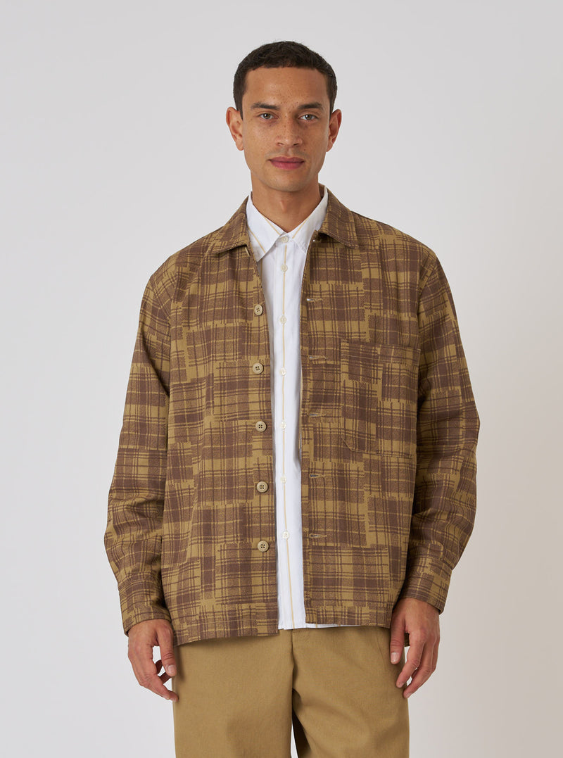 A model poses in front of a white background, wearing the Khaki Patch Check Twill Fatigue Shirt. The shirt is a brown and khaki check pattern, made of twill.