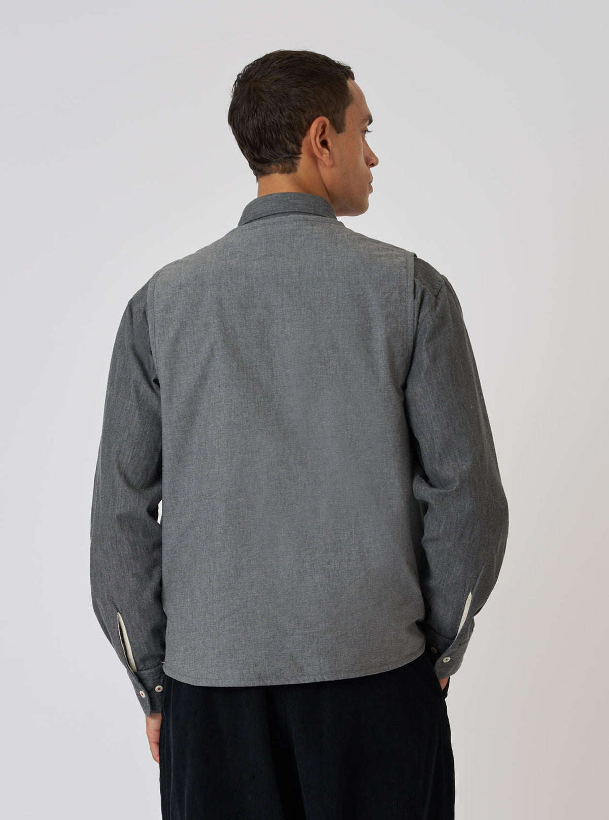 Back view of a model wearing a Charcoal Organic Chambray Parachute Liner Gilet over a long-sleeved shirt. The gilet is a charcoal color and made of organic chambray.