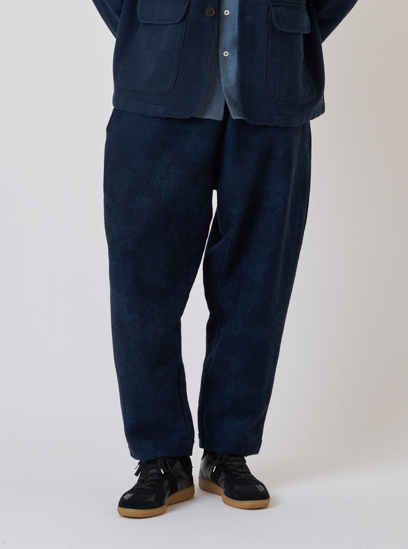 A model stands against a white backdrop wearing the Black/Indigo Denim Camo Braga Pant. The pants are a loose fit and paired with black sneakers.