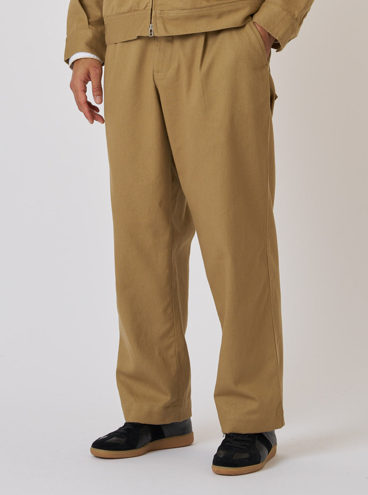 Model wears Khaki Works Twill Duke Pant in a studio shot. The khaki-colored twill pants have a relaxed fit. Worn with black sneakers. Neutral backdrop.