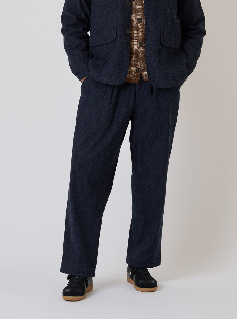 A model on a white background wears the Indigo Brushed Denim Oxford Pant. The pants are a relaxed fit and paired with a jacket and sneakers.