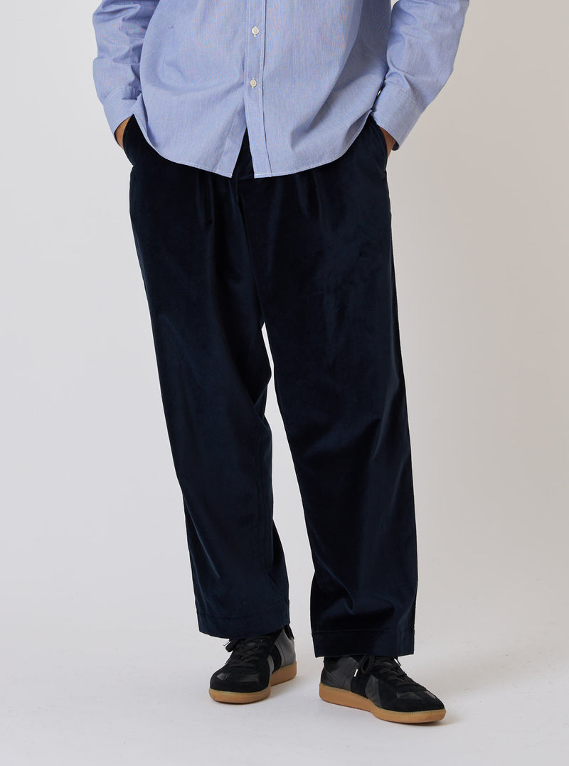A model stands against a white backdrop wearing the Navy Velveteen Oxford Pant. The pants are paired with a light blue button-down shirt and black sneakers.