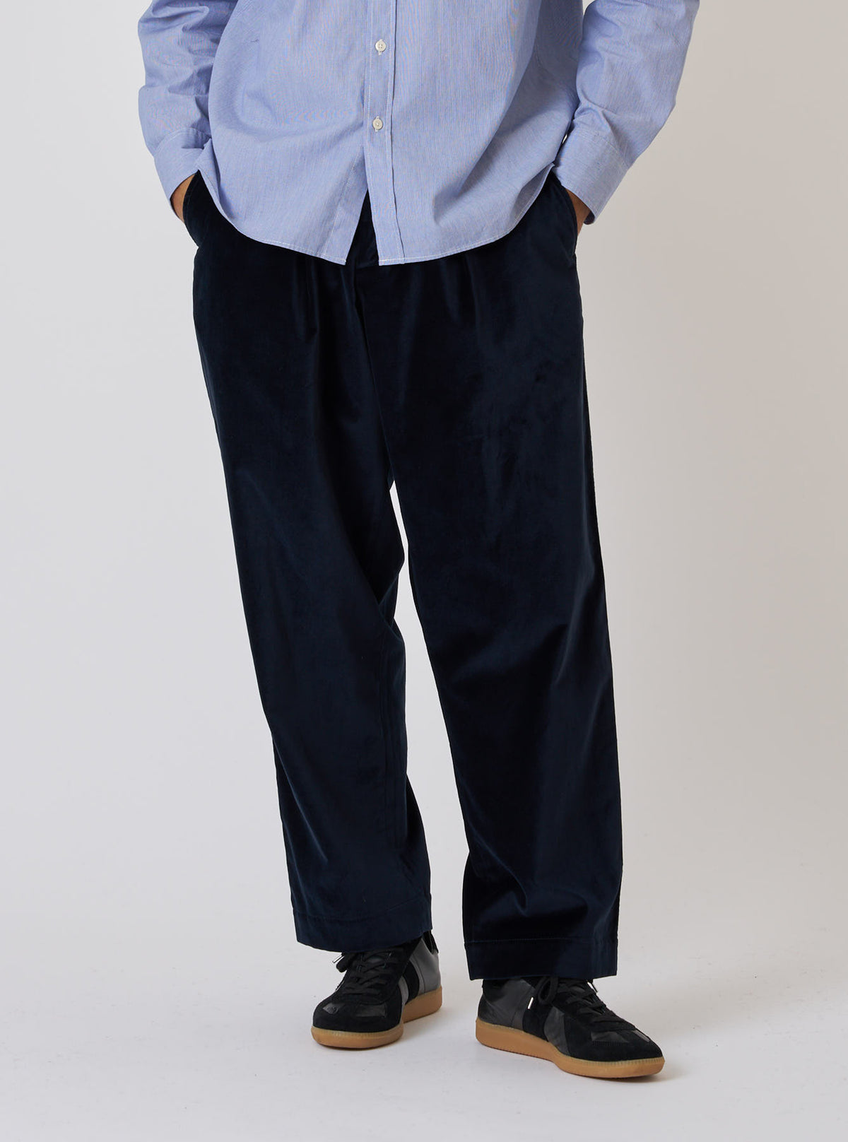 A model stands against a white backdrop wearing the Navy Velveteen Oxford Pant. The pants are paired with a light blue button-down shirt and black sneakers.