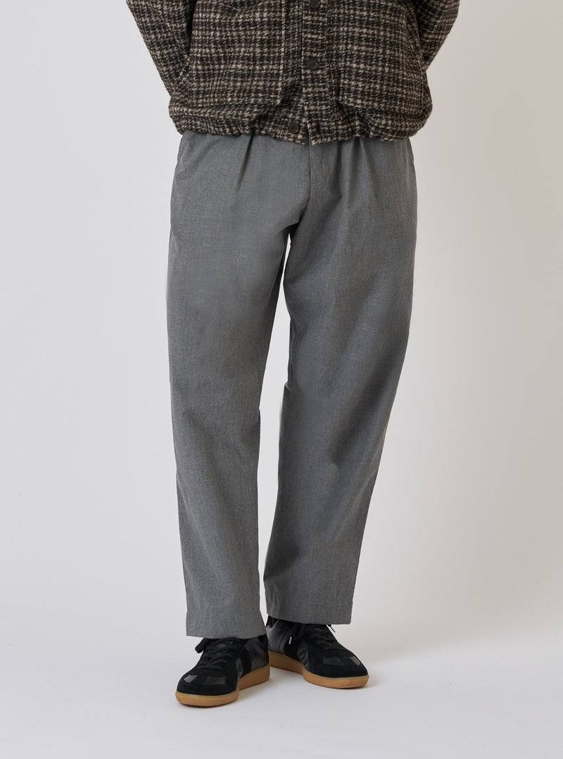 A model wears Charcoal Organic Chambray Oxford Pant in a studio setting. The pants are paired with black sneakers and a patterned jacket. The background is a clean, bright white.