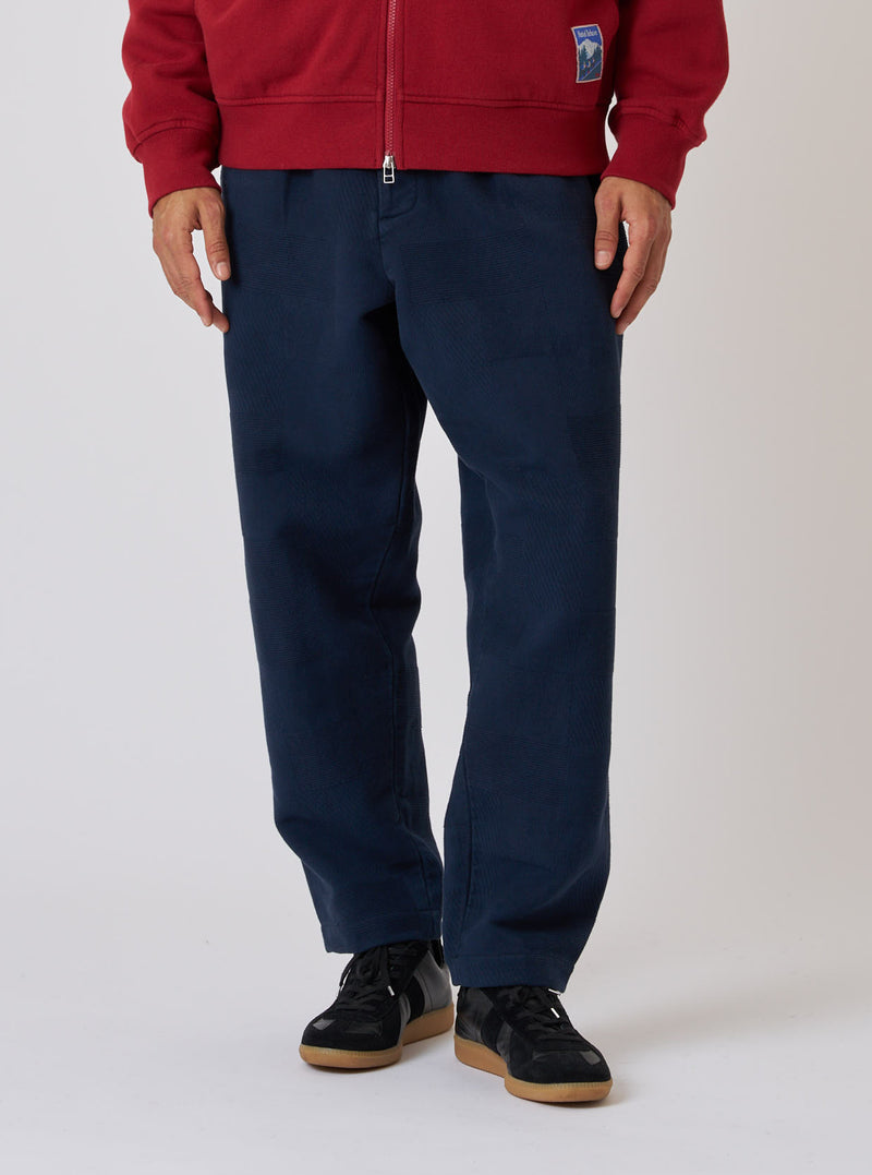 A model on a white background wears Dark Navy Patchwork Weave Cotton Pleated Track Pant. The pants are paired with black sneakers and a red jacket.