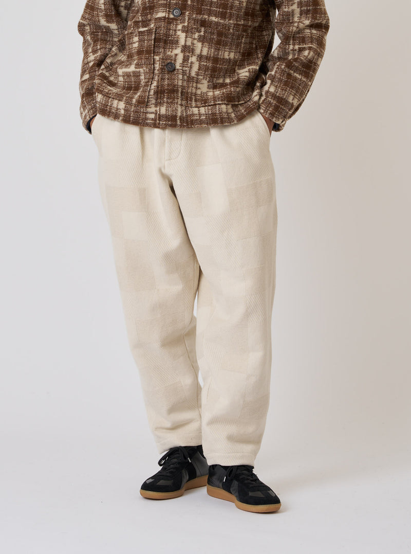 A model on a white background wears the Ecru Patchwork Weave Cotton Pleated Track Pant. The pants are ecru with a subtle patchwork pattern and a relaxed fit.