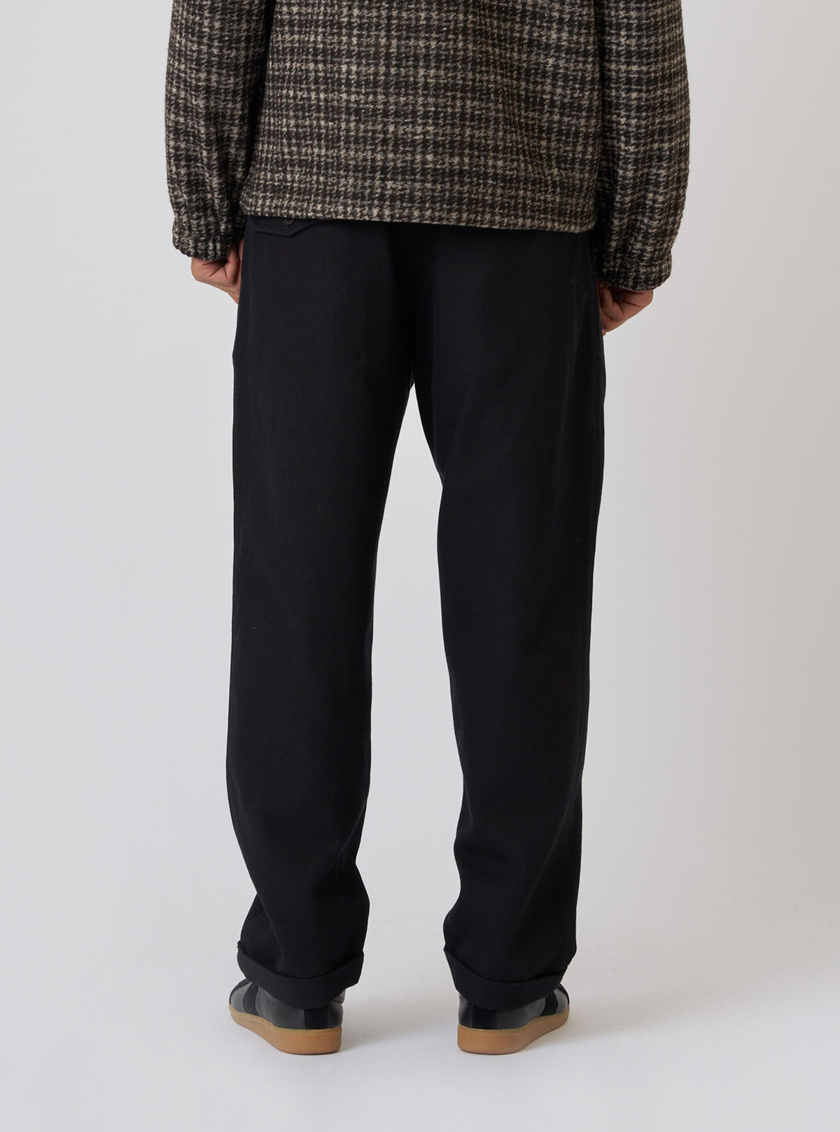 Back view of a model wearing the Black Organic Canvas Super Chino pants in a studio setting. The black chinos are paired with a patterned jacket and black sneakers.