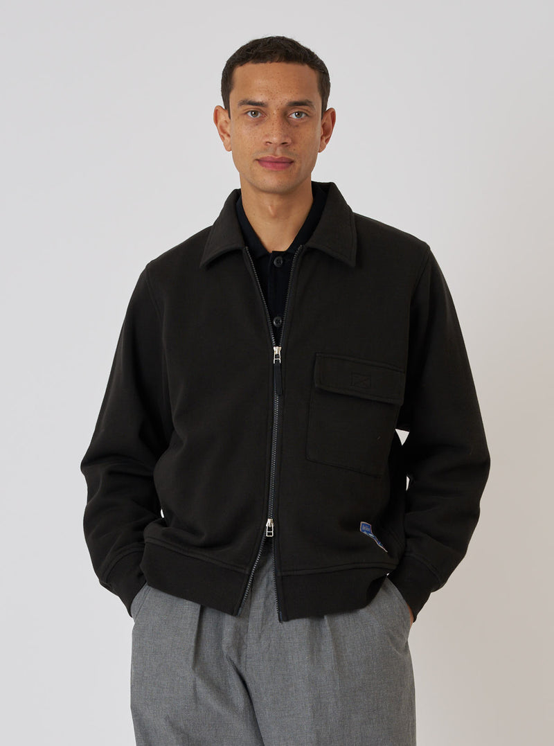 A model poses in front of a white background, wearing a Black Brush Back Sweat Tyler Sweat Jacket, a black polo shirt, and grey pants. The jacket is zipped up.