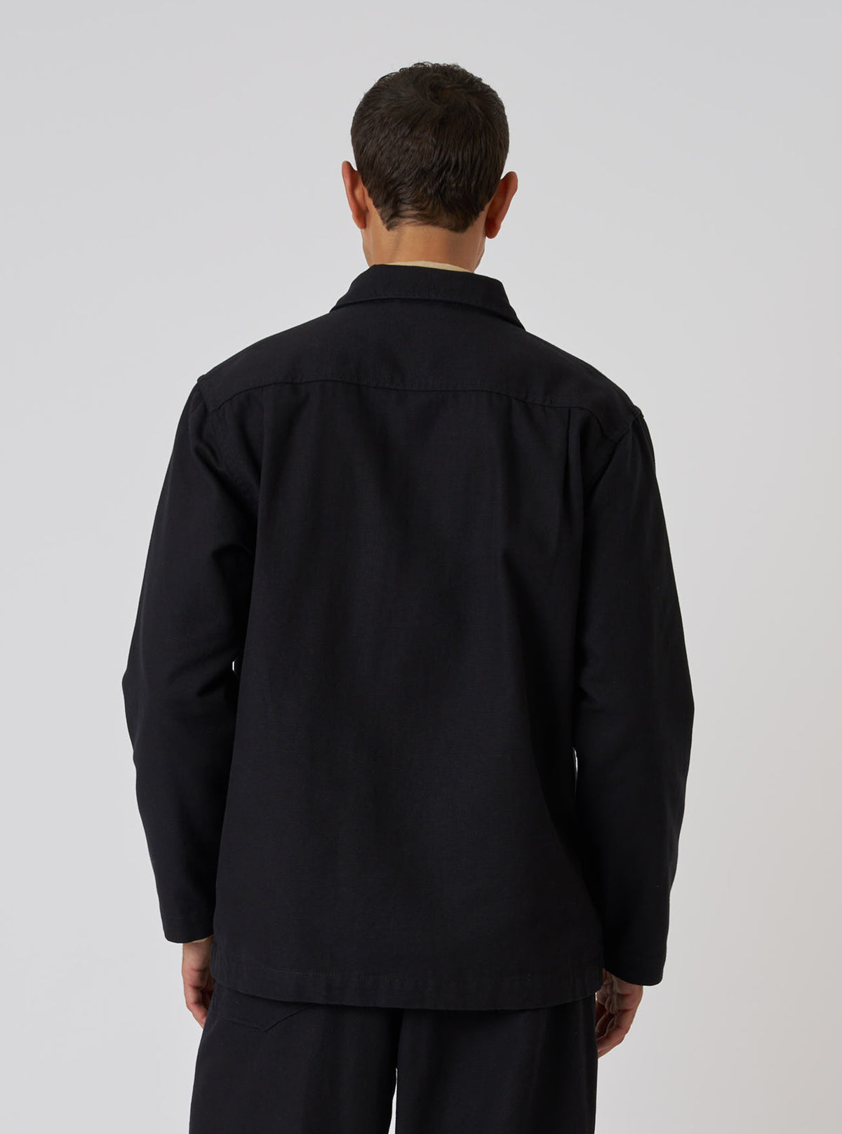 Back view of a model wearing a Black Organic Canvas Dockside Overshirt. The overshirt is made of black canvas. The background is a plain white studio backdrop.