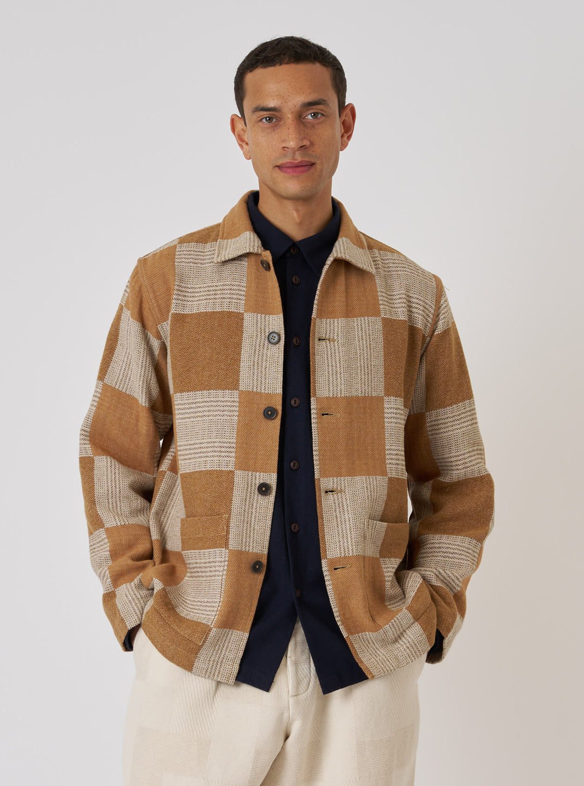 A model poses in a Sand Japanese Patchwork Easy Jacket. The jacket has a tan and cream checkerboard pattern. He wears a navy shirt and cream pants against a plain white backdrop.