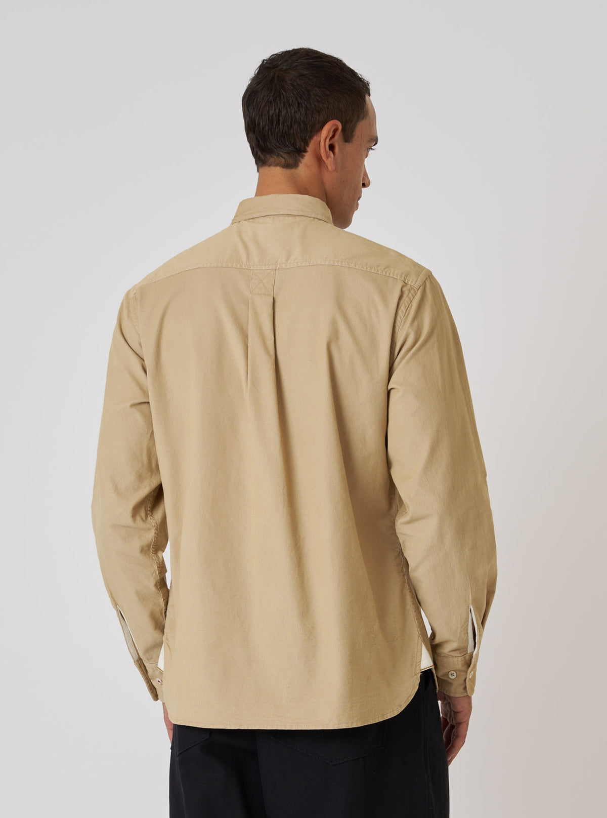 A man stands against a white backdrop, showcasing the back of the sand-colored Organic Fine Cord Daybrook Shirt. The shirt has long sleeves and a classic fit.